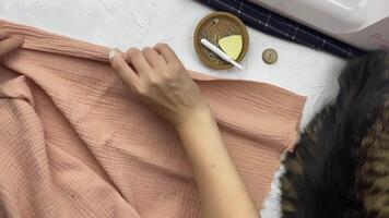 Women's hands fold the fabric and secure it with pins. Top view of the designer's workshop with a sewing machine and the back of a sleeping cat. Craft and hobby. video