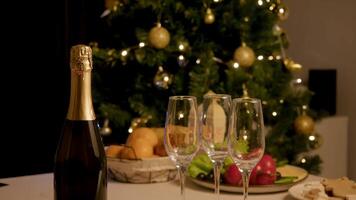 On festive table, next to several empty glasses, there is closed bottle of champagne, against backdrop of Christmas tree decorated with garlands. festive feast is ready for Christmas and New Year. video