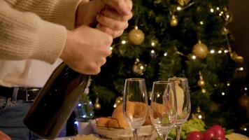 Man shakes bottle well and then, with loud pop, opens it to pour champagne into glasses with frothy head. Group of friends are celebrating Christmas or New Year around festive table. video