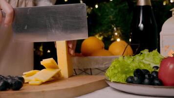 Cook cuts cheese into thin slices in order to serve it at Christmas table, which also includes variety of vegetables, fruits, and champagne. Close up video