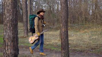 A backpacker with a guitar and a camera walks along a trail in the forest. Travel concept. Slow motion video