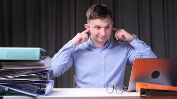 Office worker plugging ears at desk with stack of documents and laptop video