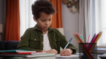Little concentrated African American boy son child kid pupil at home table studying learning writing in notebook copybook homework write distant education knowledge preparing exams learn indoors alone video