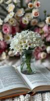 White flowers in a clear vase resting on an open book with a blurred floral background creating a serene atmosphere photo