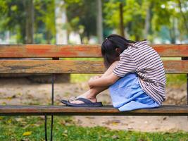 A sad little girl is sitting alone on bench in park. The little girl is unhappy and anxious because she has no one to play with. photo
