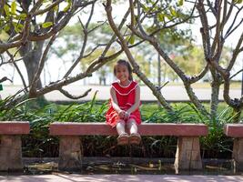 A happy little girl is sitting on a park bench. Smiling little girl with arms outstretched breathing fresh air enjoying nature in the morning. photo