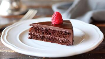 Dairy-free chocolate cake slice on a white plate topped with chocolate ganache and a fresh raspberry, photorealistic photo
