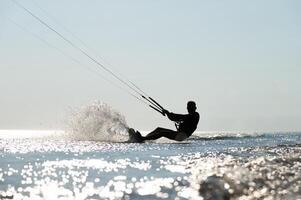 Kite surfing on waves. Silhouette of athletic man riding fast on kiteboard in sea, creating waves and splashes on water photo