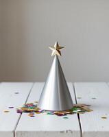 A silver party hat with a gold star on top is on a table with confetti photo