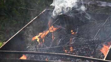 Fire burning in grill with old metal grid and messy smoldering coals and twigs covered with ashes and grey flying smoke. Flames and charcoals inside rustic mangal outdoors video