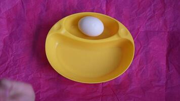 Toddler's hands break boiled egg with white spoon and peel eggshell into yellow bowl on pink rumpled paper background. Kid playing with food for breakfast video