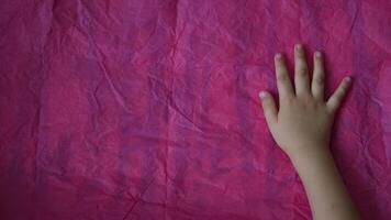 Toddler hands smooth out crumpled tissue paper on table. Directly above view of magenta wrinkled paper and kid palms of hands touch uneven pink paper texture video