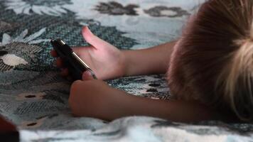 Toddler holds smartphone in hands and watches at device screen while lying in bed on blanket. Concept of kids eye vision problems due to inappropriate way to use gadgets video