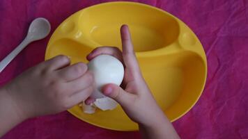 Close-up of toddler hands peel white eggshell into yellow bowl on pink crumpled paper background. Kid preparing boiled egg for breakfast. Fine motor skills development for children video