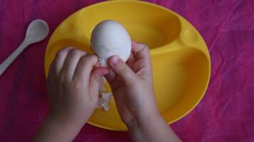 Toddler hands peel white eggshell into yellow bowl on pink crumpled paper background. Kid preparing boiled egg for breakfast. Fine motor skills child development video
