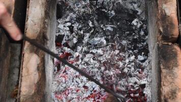 Hand holding iron poker and shake red smoldering coals with red sparks and blur grey ash flying over. Putting grill grid with defocus traditional sausages Bratwurst atop embers in barbecue brazier video