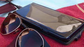 Blur reflections in black mirrored surface of smartphone and sunglasses. Defocus reflection of white beach umbrella in cell phone screen. Mobile phone with charging cable. Summer vacation concept video