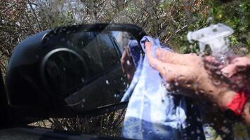 Defocused view through unclean glass window in process of wet car wash with hands and detergent foam spray. Male hands clean up glass surface with rag video