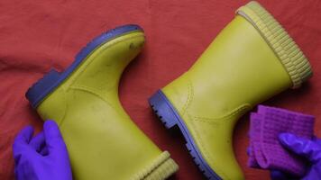 Hands in purple rubber gloves clean and wipe dirty pair of kids yellow boots with wet rag. Top view on pair of waterproof rubber boots. Housework concept video