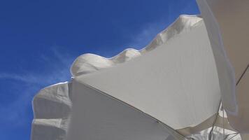 Fluttering frills of white textile umbrellas against blue sky background with copy space. View from under parasol on beach. Seaside relaxation concept video