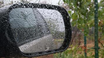 Defocus side mirror of car with blurred reflection through raindrops dots on glass window surface. Looking out from wet car window in drizzle with bokeh background video