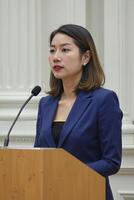 A photograph of an Asian woman with shoulder-length brown hair, wearing a blue blazer and black top, speaking at a podium with a microphone. photo