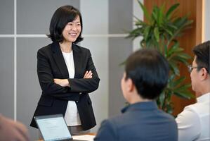 A photograph of a confident Asian woman in a black suit, standing with arms crossed, in a modern office with other professionals in the background. photo