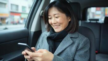 A middle aged Asian woman with shoulder length black hair, wearing a gray coat and scarf, smiles while using a smartphone in a car photo