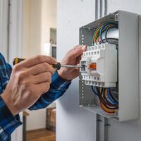Electrician tightens component on white electrical box with yellow screwdriver, wires visible For Social Media Post Size photo