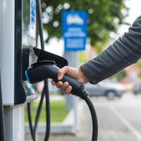 Hand holds black EV charging nozzle at wall mounted station, blue sign blurred For Social Media Post Size photo