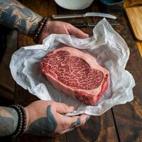 Tattooed hands hold beef in parchment paper, marbling pattern visible on meat For Social Media Post Size photo