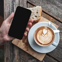 Hand presents smartphone next to cappuccino with latte art, rustic table and bread For Social Media Post Size photo