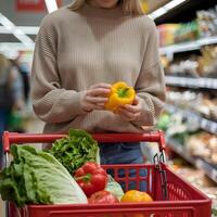 Woman in beige sweater holds yellow bell pepper, red cart, blurred grocery store background For Social Media Post Size photo