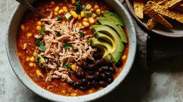 Comforting chicken soup with corn and beans, topped with avocado and served with chips photo