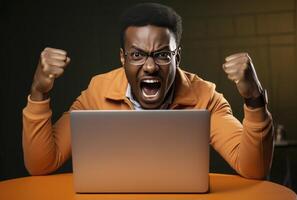 A dark skinned man with glasses sits at an orange table, furiously yelling at a laptop photo