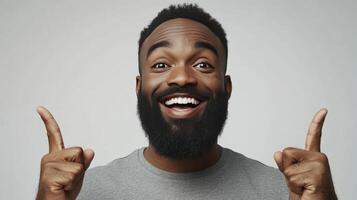 A portrait of a smiling Black man with a beard, pointing upwards with both index fingers against a plain grey background photo