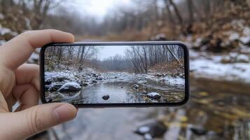 A hand holds a smartphone capturing a winter stream scene. Snow covers the banks, trees are bare, and the water is calm photo