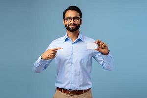 Smiling bearded handsome Indian man holding empty, white business card, pointing finger on mockup photo