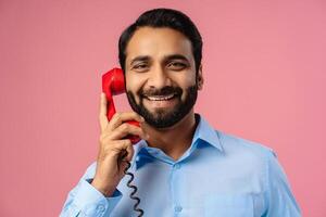 Portrait of smiling, handsome Indian man holding red phone, answering call, looking at camera photo