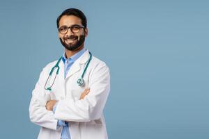 Smiling, Indian doctor wearing white coat and stethoscope and stylish eyeglasses with crossed arms photo