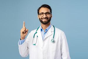 Portrait of handsome, smiling, Indian man wearing white coat, stylish eyeglasses pointing finger up photo