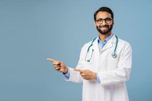 Smiling Indian man, doctor wearing white coat, stylish eyeglasses, pointing finger on copy space photo