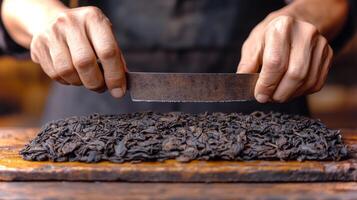 Skilled hands maneuver a sharp knife to slice through a dense block of aged tea leaves, revealing their rich texture under warm afternoon light in a workshop setting. photo