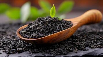 A wooden spoon filled with loose black tea leaves sits on a slate surface. A small green tea shoot emerges from the pile, adding a touch of freshness. photo
