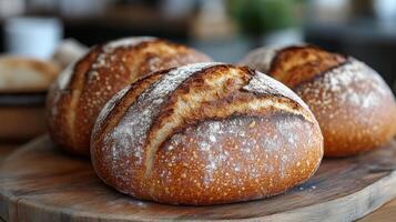 Three round artisan bread loaves are arranged on a wooden board, showcasing a crusty, golden-brown exterior with a dusting of flour. The cozy kitchen ambiance enhances their appeal. photo