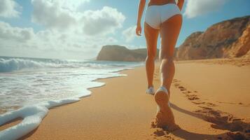 A woman strolls barefoot along the shoreline, leaving footprints in the wet sand as gentle waves lap at her ankles. The bright sun illuminates the scenic beach. photo