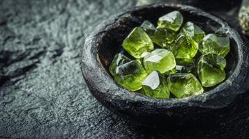 Bright green gemstones are carefully placed in a deep bowl resting on a rough, dark stone surface. The arrangement showcases the unique shapes and transparent qualities of each stone. photo