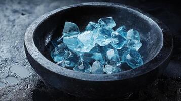 A collection of sparkling blue gemstones rests in a black bowl on a textured surface, showcasing their vibrant hues in a low light setting. The arrangement highlights the stones' clarity and facets. photo