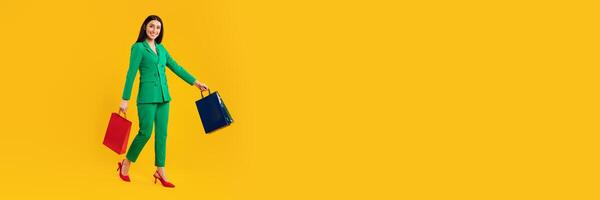 A joyful woman in a bright green suit and red heels walks while holding two shopping bags. The background is a vibrant yellow, creating a lively atmosphere perfect for Black Friday shopping. photo
