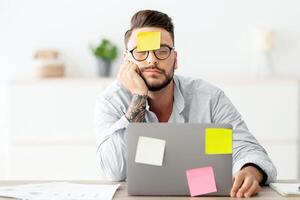 A lot of work concept. Tired freelancer man sleeping at workplace with stickers on foehead and computer, working on laptop at home office, sitting at table in living room, free space photo